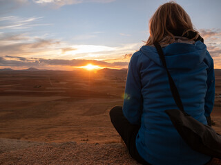BLONDE WOMAN WITH BLUE JACKET AND TRIPOD HANGED ON THE BACK, SITTING WATCHING THE SUNSET ON A HILL IN CONSUEGRA, SPAIN