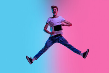 Fototapeta na wymiar Showing tablet screen. Young caucasian man's jumping high on gradient blue-pink studio background in neon light. Concept of youth, human emotions, facial expression, sales, ad. Beautiful model in