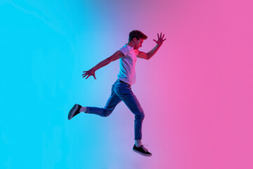 Obraz na płótnie Canvas Running on. Young caucasian man's jumping high on gradient blue-pink studio background in neon light. Concept of youth, human emotions, facial expression, sales, ad. Beautiful model in casual.