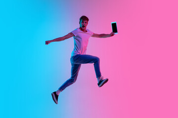 Fototapeta na wymiar Winner with tablet. Young caucasian man's jumping high on gradient blue-pink studio background in neon light. Concept of youth, human emotions, facial expression, sales, ad. Beautiful model in casual.