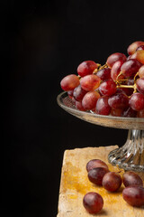 View of half glass fruit bowl with wet red grapes, selective focus, on wooden table, vertical black background