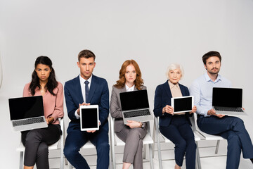 Multiethnic businesspeople showing devices with blank screen while sitting on chairs on grey background