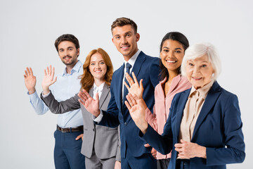 Smiling businessman waving hand at camera near multicultural colleagues on blurred foreground isolated on grey