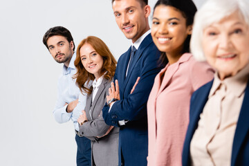 Fototapeta premium Smiling businesspeople with crossed arms looking at camera near multiethnic colleagues on blurred foreground isolated on grey
