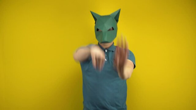 Young man in a cardboard jackal mask dances on a yellow background. Man in a green polo and mask.
