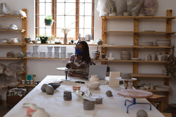 Mixed race female potter in face mask working in pottery studio