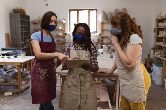 Two caucasian and one mixed race female potters in face mask working in pottery studio