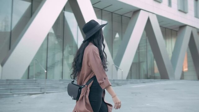 Fashionable brunette young woman wearing a pink coat and a black hat walking to the right on the sidewalk. Slow motion, 4k, tracking shot, side view, cinematic