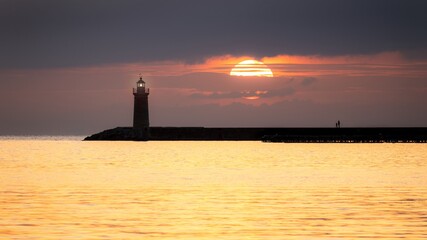 Fototapeta premium Lighthouse at sunset with large sun, golden sea and people, Andratx, Mallorca, Spain.