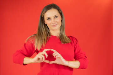 Portrait of young, pretty woman in casual sweater showing with her finger heart figure, sending love to her lover, standing over red background