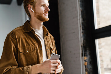 Focused redhead guy in earphones using cellphone and looking at window