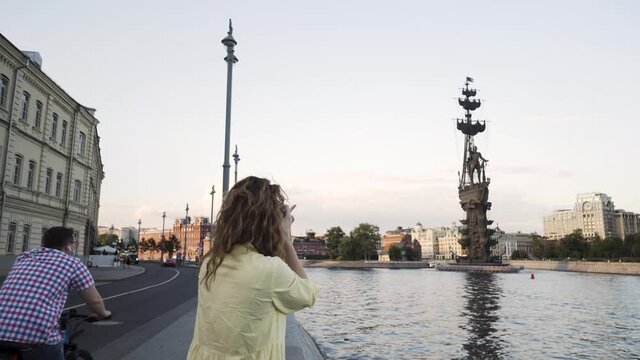 Woman with a professinal camera taking pictures of Neva river in Saint Petersburg, Russia. Action. Female tourist with curly hair photographing the ship monument in the name of Peter the First.