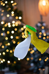 A hand in a yellow glove holds a bottle of detergent on the background of New Year's lights. General professional cleaning before Christmas. Spray disinfection, safety