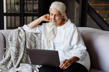 Retired senior woman sitting at home using laptopn a couch