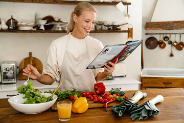 Beautiful joyful woman reading cookbook and smiling while making lunch