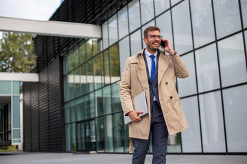 Portrait of a smiling businessman talking on the phone with a laptop in his hand in front of office building.