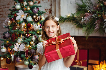Christmas, holidays and childhood concept-a smiling girl with a gift box at home near a decorated Christmas tree.