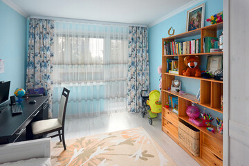 Cozy bright children's room with blue walls pastel carpet on gray wooden floor wooden bookshelf bed and desk with globe and computer. Real estate apartment.