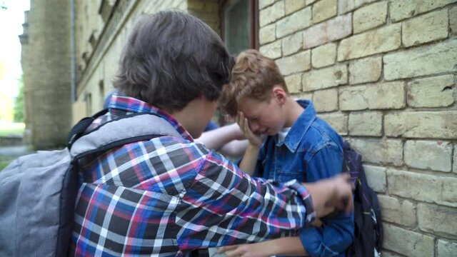Aggressive classmates bullying frightened redhead nerd on schoolyard. Side view portrait of violent Caucasian schoolboys teasing scared boy outdoors. Childhood and aggression concept.