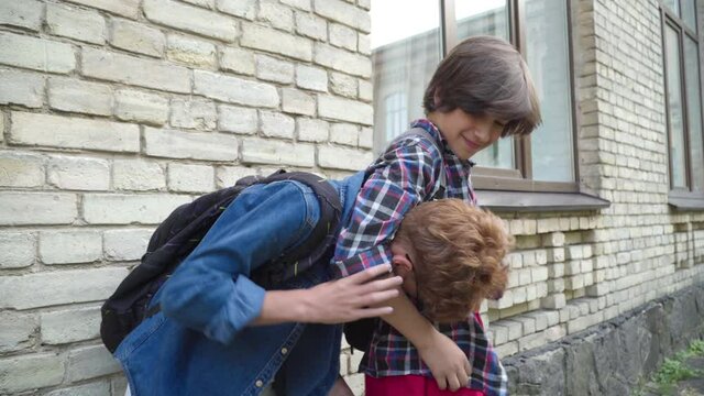 Side view of violent brunette schoolboy teasing redhead nerd in eyeglasses outdoors. Aggressive Caucasian boy rubbing head of victim on schoolyard during break. Bullying and lifestyle.