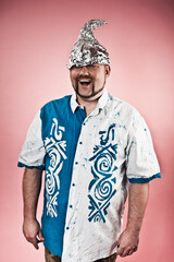 Studio photo of a goofy looking man wearing a tin foil hat on dark pink background.