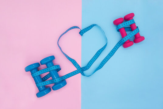 Composition with blue and pink dumbbells and a tailor's centimeter in the form of a heart on a pink-blue background. Sports activities for two. Valentines day concept in fitness club.