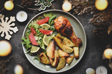 Winter festive food concept. Top view of dark vintage background, garland and candles. Christmas and New Year background. Baked chicken legs and potatoes.