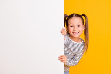 Photo of young happy smiling little girl child kid stand behind white wall isolated on yellow color...
