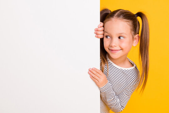 Naklejki Portrait of young happy smiling curious little girl child kid behind white wall banner look in copyspace isolated on yellow color background