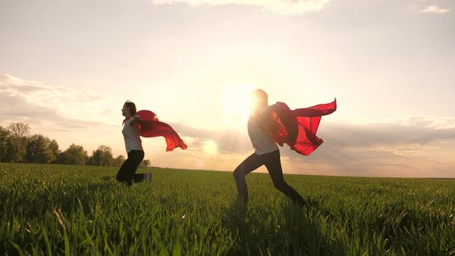 happy girls play superheroes they run across green field in red cloak, cloak flutters in wind. childrens games and dreams. Slow motion. teenager dreams of becoming superhero. young girls in red cloak