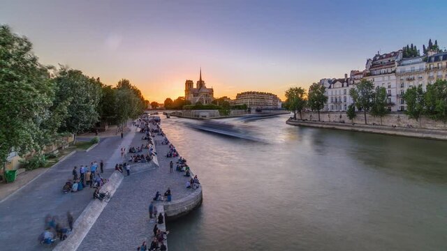 Rear view of Notre Dame De Paris cathedral at sunset with sun in the frame timelapse. View from Tournelle bridge. People sitting on waterfront. Orange sky in background. Paris, France, Europe.