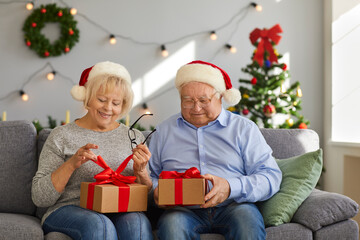 Obraz na płótnie Canvas Happy aged couple in Christmas festive hats sitting on sofa and unpacking holiday presents