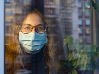 Depressed young girl wearing face mask looking out of window standing at home during coronavirus self-isolation. Social distancing, quarantine. Patient isolated in house to prevent infection.