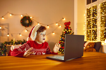 Fototapeta na wymiar Shocked child sitting at table with laptop looking at real Santa Claus on video chat