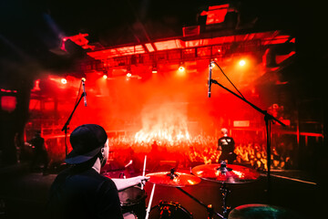 Band on stage. Drummer playing drum set at concert on stage. Music show. Bright scene lighting in club, drum sticks in hands. Fans burn red flares at a rock concert. View from the stage to the drummer