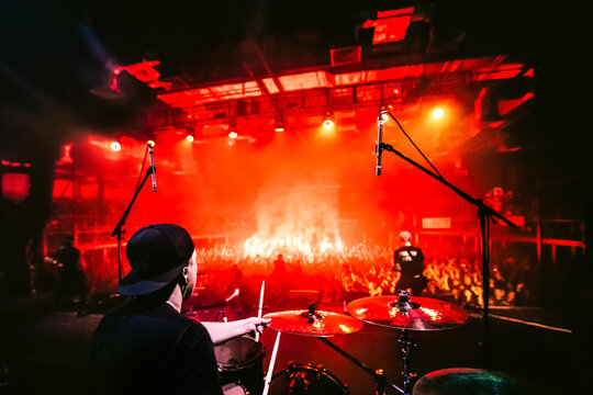 Band On Stage. Drummer Playing Drum Set At Concert On Stage. Music Show. Bright Scene Lighting In Club, Drum Sticks In Hands. Fans Burn Red Flares At A Rock Concert. View From The Stage To The Drummer