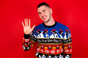 Young handsome Caucasian man wearing Christmas sweater against red wall, Waiving saying hello happy and smiling, friendly welcome gesture.