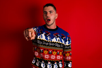 Young handsome Caucasian man wearing Christmas sweater against red wall, Pointing with finger surprised ahead, open mouth amazed expression, something on the front.
