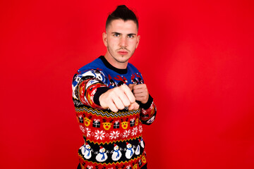 Young handsome Caucasian man wearing Christmas sweater against red wall, Punching fist to fight, aggressive and angry attack, threat and violence