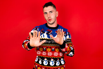 Young handsome Caucasian man wearing Christmas sweater against red wall, Moving away hands palms showing refusal and denial with afraid and disgusting expression. Stop and forbidden.