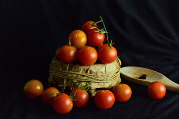 Fototapeta na wymiar Dark background. Rustic bowl with cherrys tomatoes and more scattered on black cloth, light entering from the side, together also a wooden spoon with a heart cutout.