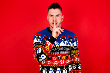 Young handsome Caucasian man wearing Christmas sweater against red wall,  makes silence gesture, keeps finger over lips. Silence and secret concept.