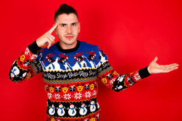 Obraz premium Young handsome Caucasian man wearing Christmas sweater against red wall, confused and annoyed with open palm showing copy space and pointing finger to forehead. Think about it.