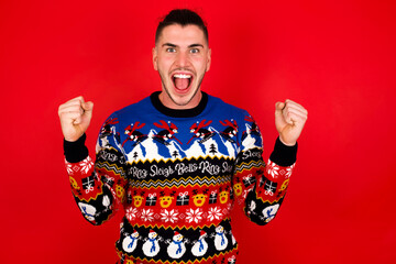Young handsome Caucasian man wearing Christmas sweater against red wall,  celebrating surprised and amazed for success with arms raised and open eyes. Winner concept.
