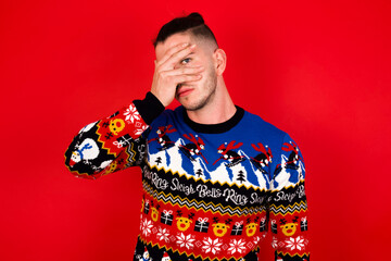 Obraz premium Young handsome Caucasian man wearing Christmas sweater against red wall, peeking in shock covering face and eyes with hand, looking through fingers with embarrassed expression.
