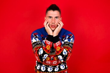 Stupefied Young handsome Caucasian man wearing Christmas sweater against red wall,  expresses excitement and thrill, keeps jaw dropped, hands on cheeks, has eyes popped out