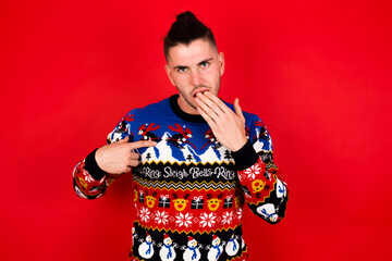 Shocked Young handsome Caucasian man wearing Christmas sweater against red wall,  look surprisedly down, indicates at blank  with fore finger, Scared model