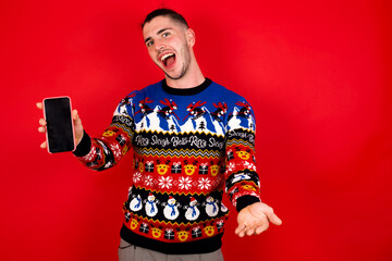 Young handsome Caucasian man wearing Christmas sweater against red wall,  with a mobile. presenting smartphone
