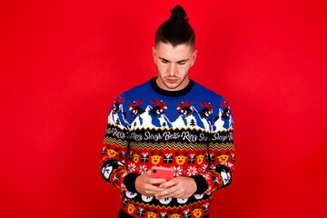 Excited Young handsome Caucasian man wearing Christmas sweater against red wall,   winking and eye hold smart phone use read social network news