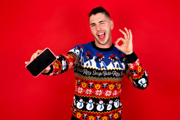 Young handsome Caucasian man wearing Christmas sweater against red wall,  holding in hands cell showing ok-sign